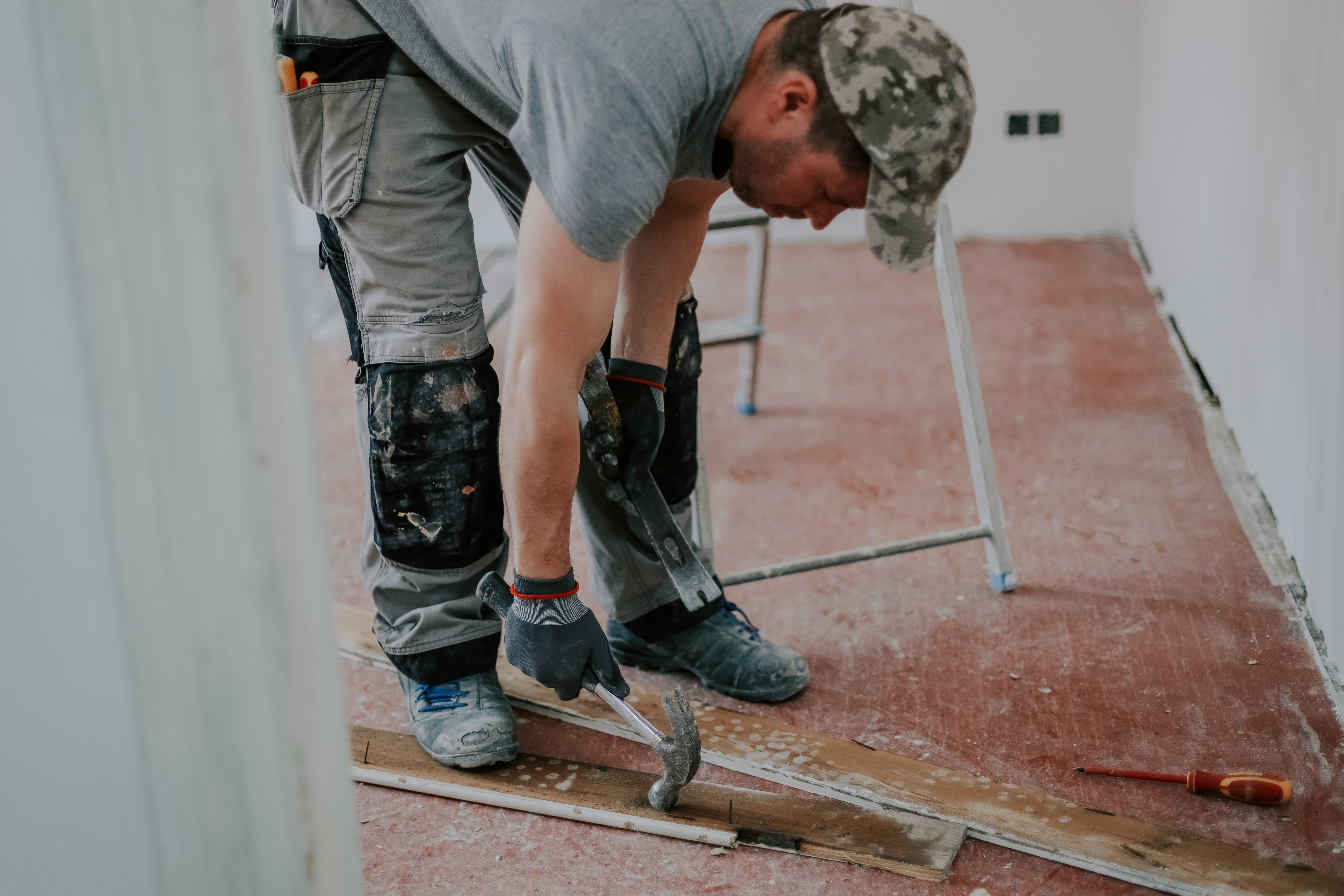 Young handsome caucasian man in uniform and textile gray gloves pulls out nails with a hammer and crowbar from a dismantled old wooden plinth while standing in an inclination, close-up side view with selective focus.Construction work concept.