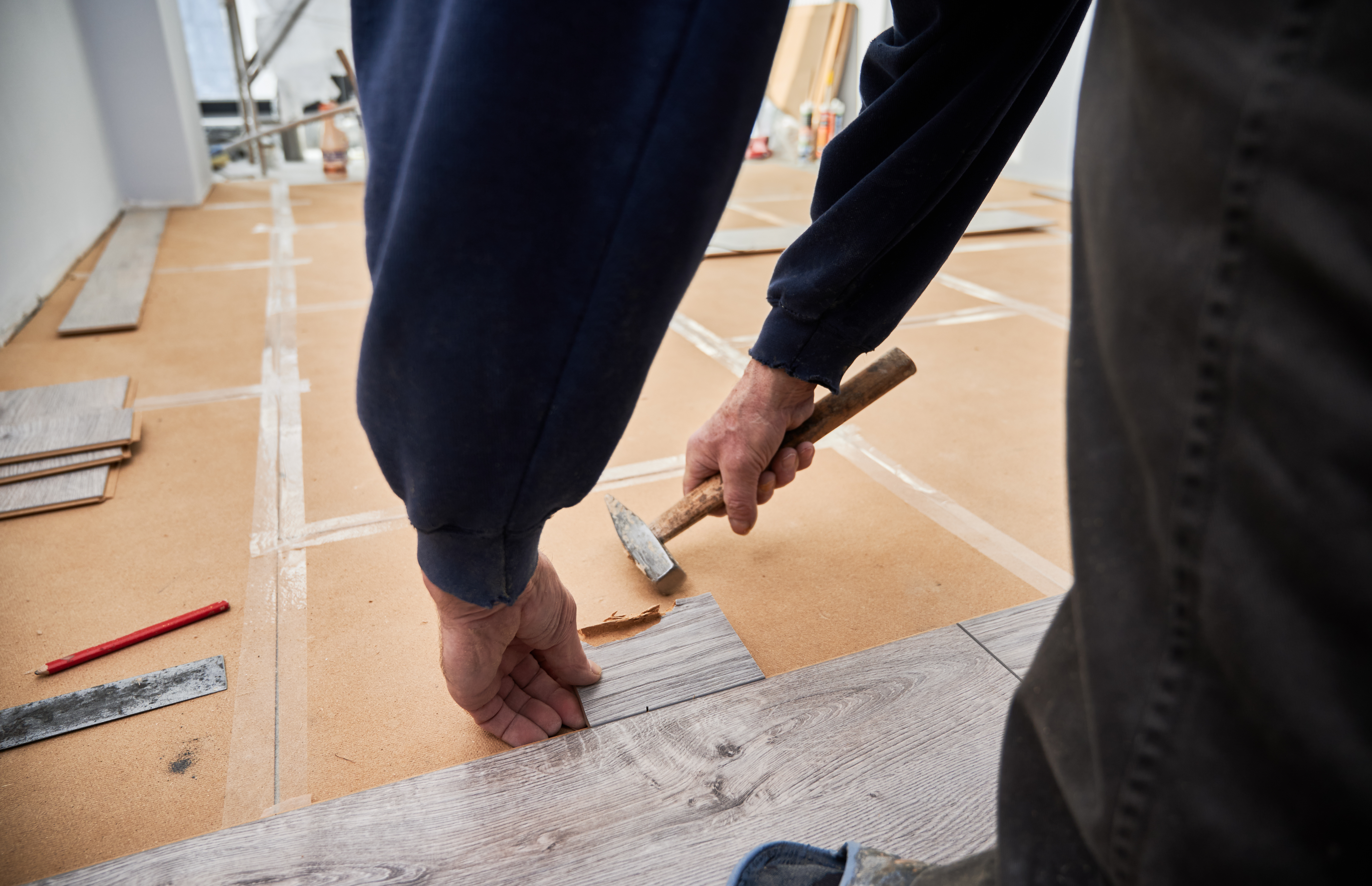 Close up of man construction worker using hammer tool while installing laminate flooring in apartment under renovation. Male hands laying laminate wooden panel on the floor. Home renovation concept.